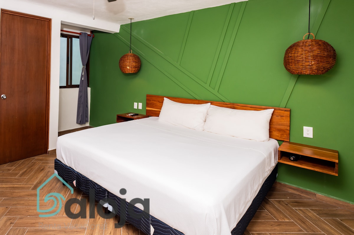 A spacious sleeping area is presented with a large bed adorned with crisp white linens. The green accent wall adds a refreshing touch, complemented by woven pendant lights on either side. Natural light filters through a nearby window, enhancing the inviting atmosphere.