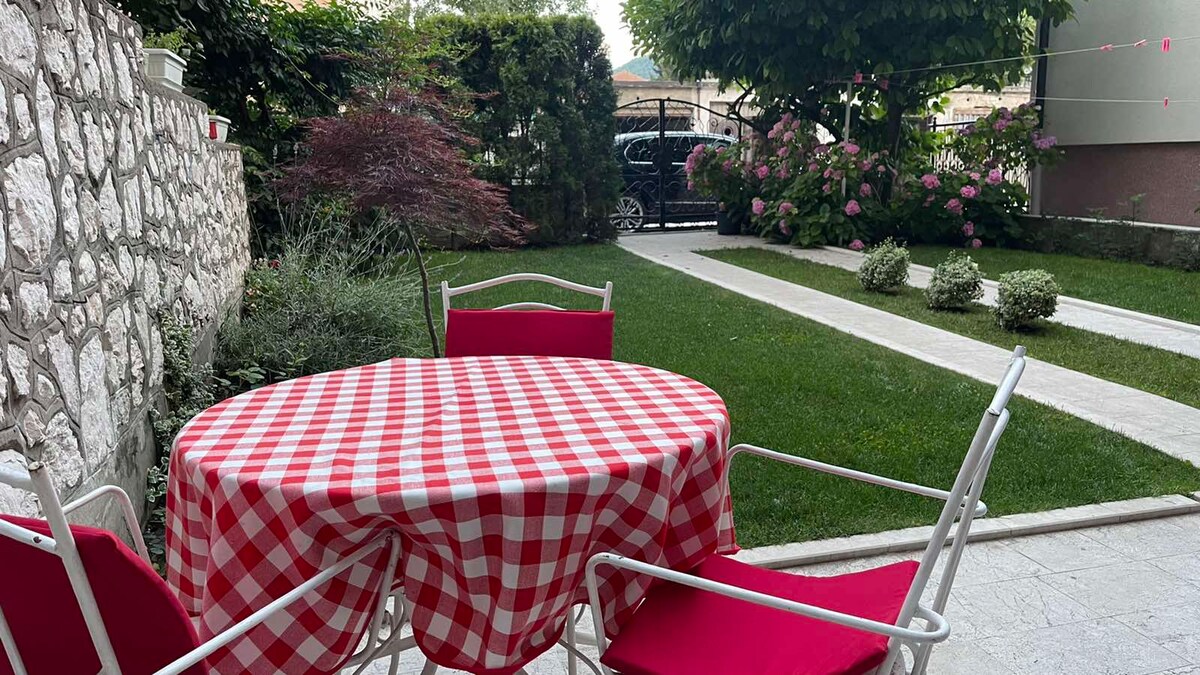 An outdoor space features a round dining table draped with a red and white checkered tablecloth, surrounded by four white chairs with red cushions. Lush green grass and decorative shrubs create a serene garden atmosphere, complimented by flowering plants in the background.