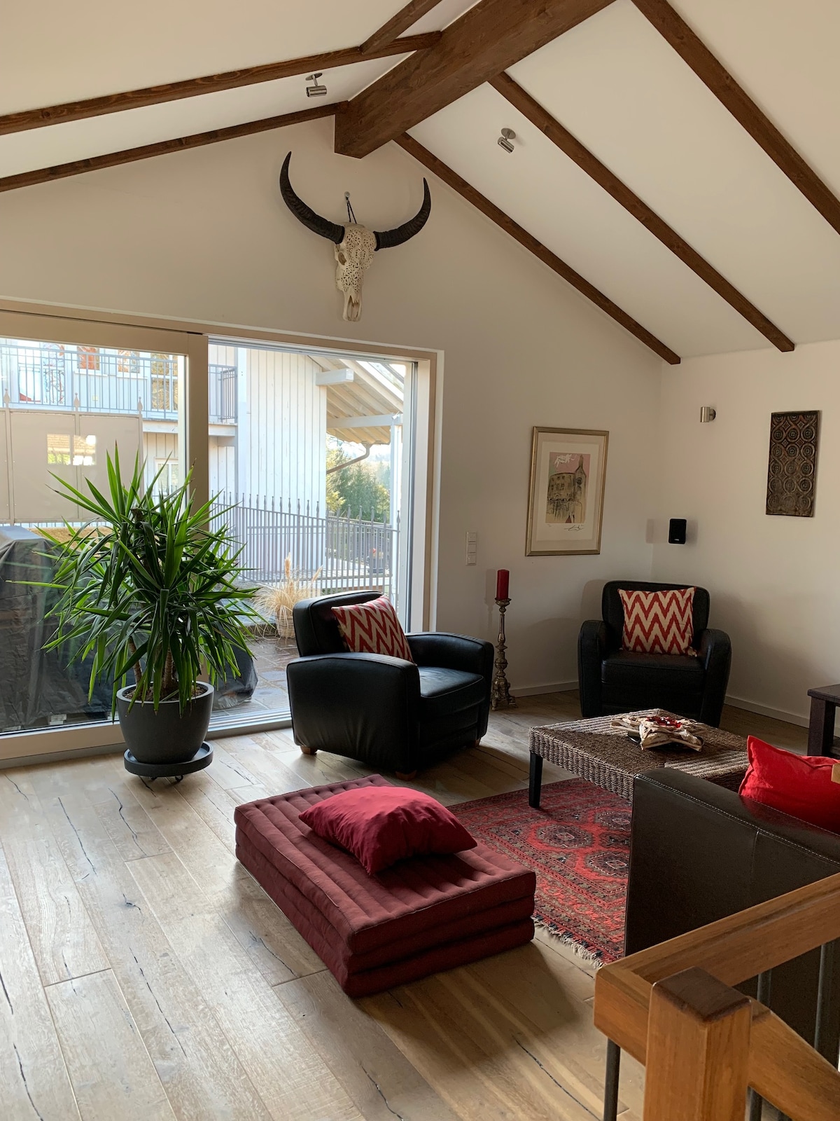 A cozy living space features a potted plant beside two black chairs with patterned cushions. A wooden coffee table sits on a patterned rug, with a crimson cushion positioned on a low bench. Large glass doors open to an outdoor area, and decorative items adorn the walls.