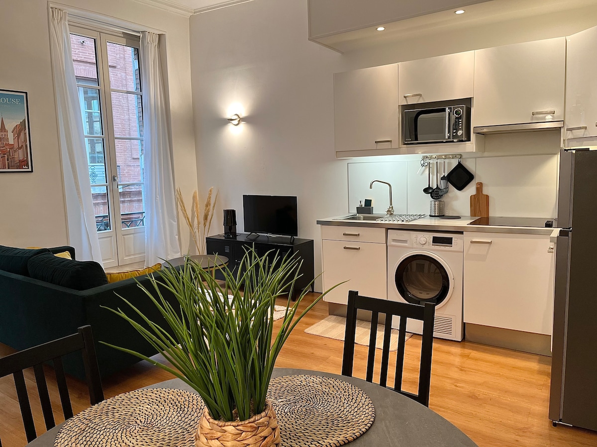 A modern kitchen area equipped with essential appliances, including a microwave and refrigerator, is visible. A sleek black television is positioned near a cozy green sofa. A circular dining table, adorned with a decorative plant, complements the warm wooden flooring and ample natural light from the windows.