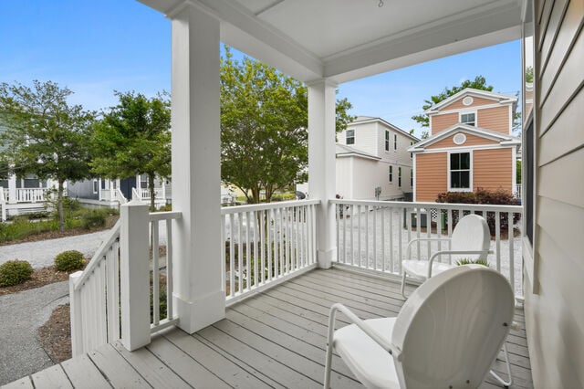 A welcoming porch area features two white chairs positioned beside a small potted plant. The view showcases a neatly landscaped pathway and adjacent homes, framed by greenery and a clear blue sky.