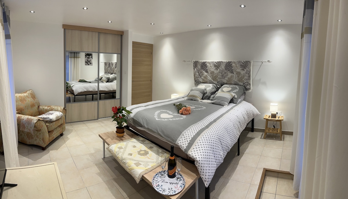 A spacious bedroom features a large bed adorned with soft linens. A cozy chair is placed nearby, with warm lighting from bedside tables. A mirrored wardrobe reflects the contemporary decor, contributing to an airy feel, while a small table showcases decorative items and a beverage.