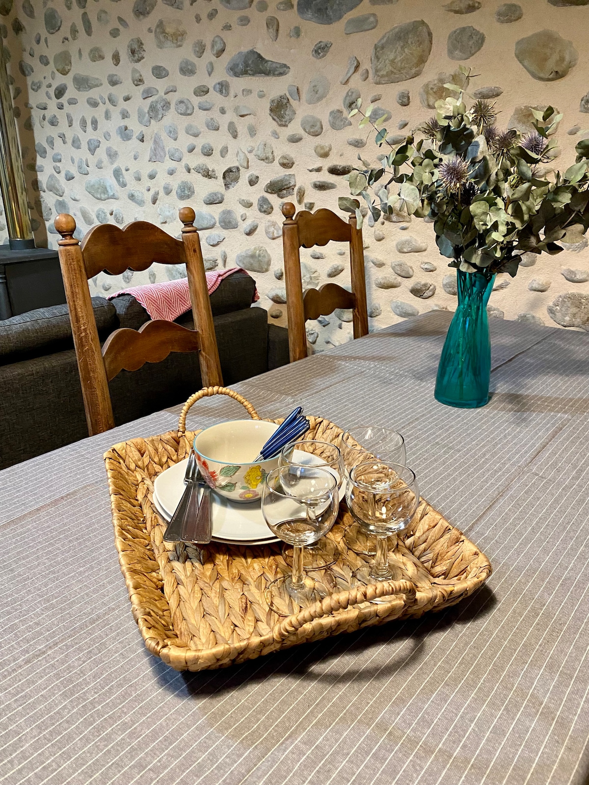 A woven tray is placed on a textured tablecloth, holding a bowl, utensils, and several glasses. Behind, wooden chairs with a rustic design are arranged around the table, while a vase of dried flowers adds a decorative touch. The stone wall provides a natural backdrop.
