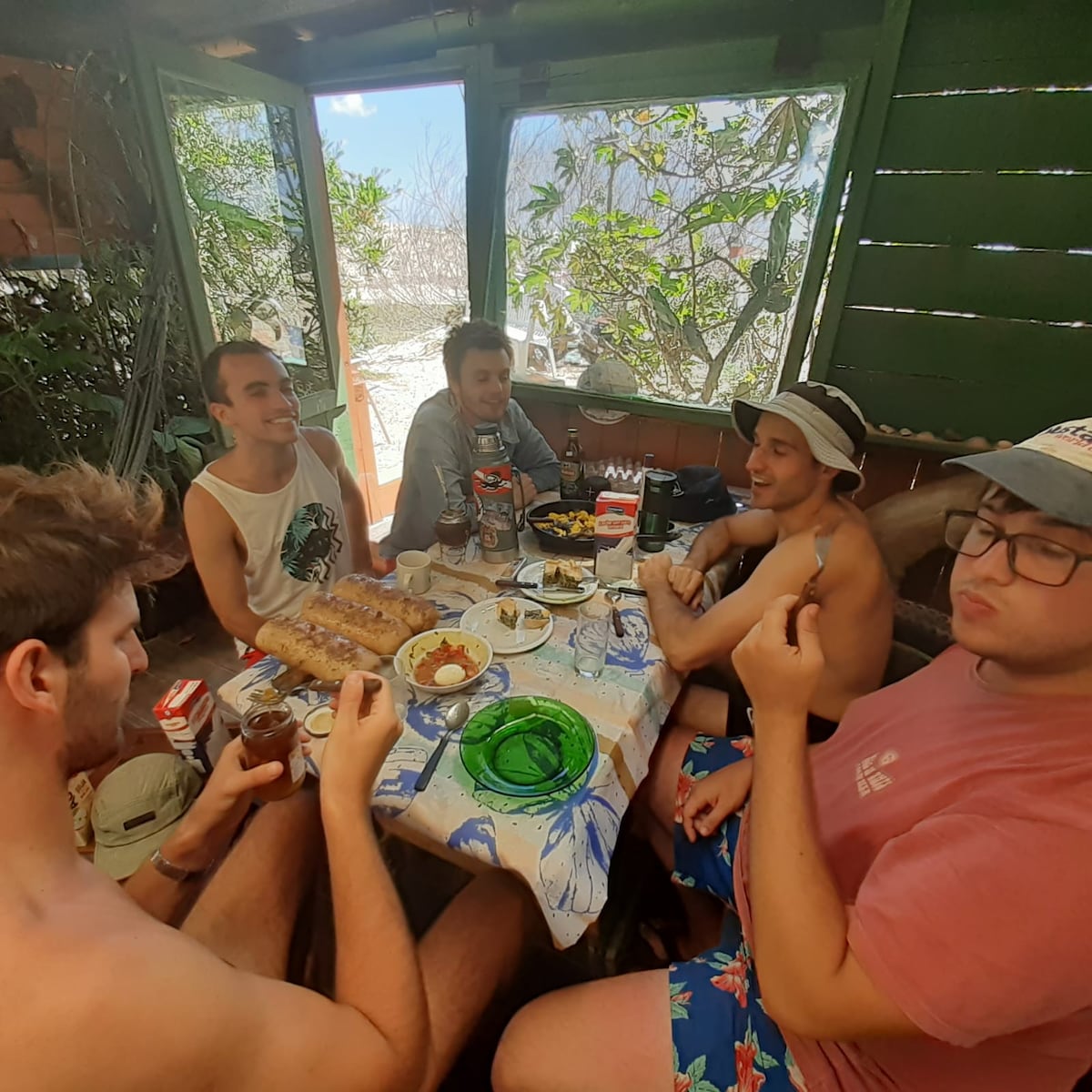 A gathering is shown around a table set outdoors, surrounded by windows providing a view of greenery. Various dishes are visible on the table, with people casually engaged in conversation. The atmosphere reflects an informal meal setting in a natural environment.