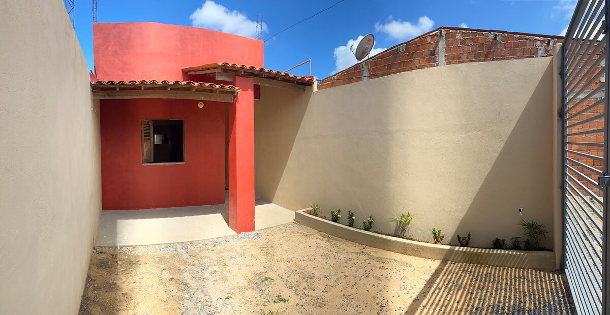 A private outdoor area is framed by simple walls, featuring a red accent wall and a small window. The space is covered with light gravel and includes a few low-growing plants arranged along the perimeter. A satellite dish is visible above the roof.