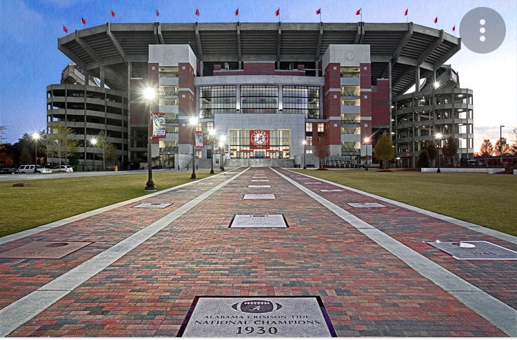 The image displays the exterior of a stadium, prominently featuring brick walkways that lead toward the main entrance. Tall lights illuminate the pathway, while banners and flags adorn the structure, signifying a prominent sports venue. Surrounding greenery frames the view.