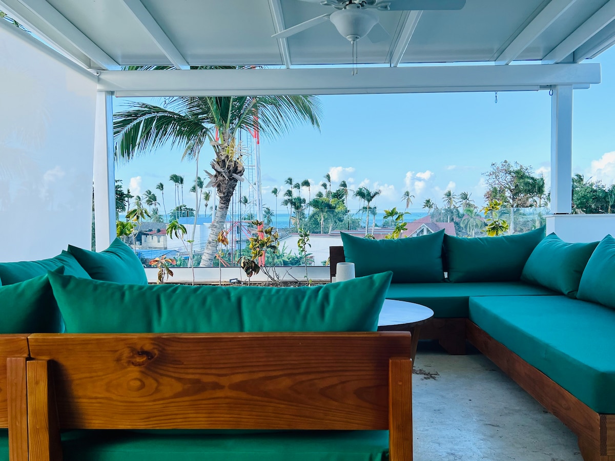 A spacious terrace features a cozy seating area with green cushions against a wooden frame. Tropical plants and palm trees can be seen in the background, with a distant view of the sea and sky, creating a serene outdoor setting.