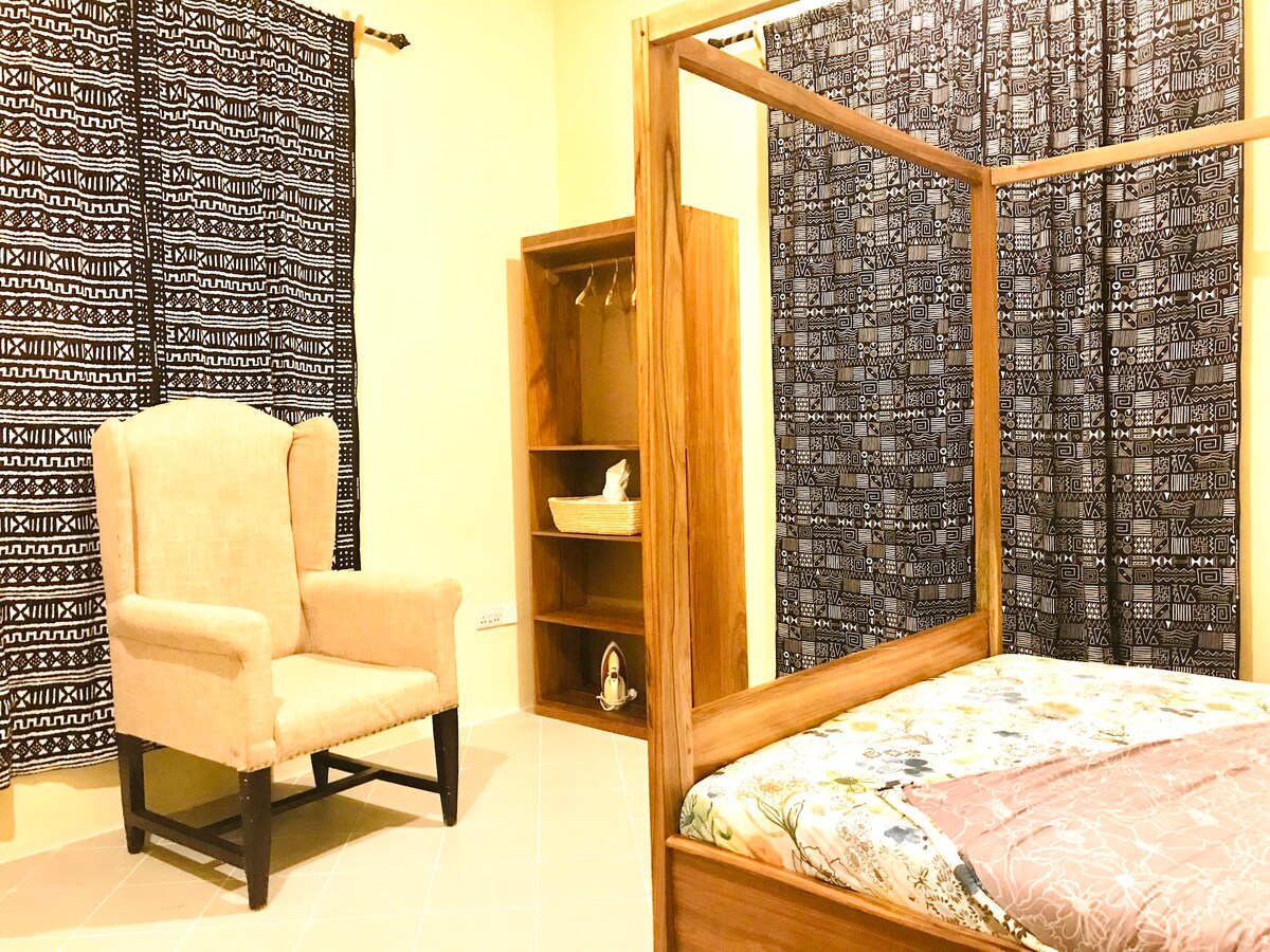 A spacious room features a wooden four-poster bed with a colorful bedding set. A comfortable armchair is positioned nearby, accompanied by a wooden shelving unit. The walls are adorned with dark patterned curtains, enhancing the room's contemporary aesthetic.