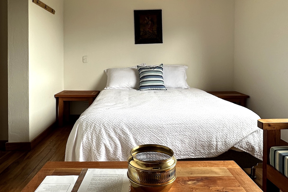 غرفة بسرير مزدوج وطاولتين في الليل وشماعة خشبية ريفية وأريكة مع وسائد مخططة وديكور بسيط وإضاءة طبيعية. أسلوب دافئ وعملي ومرحّب.