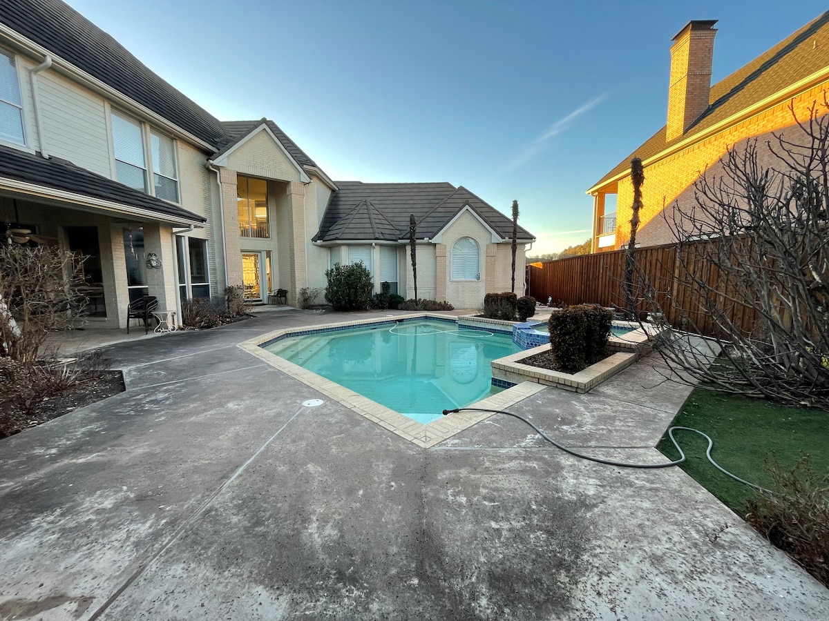 A spacious outdoor area features a swimming pool surrounded by a concrete patio. Lush shrubbery is present along the perimeter, with a smooth lawn area visible. Two homes are located alongside the pool, providing a welcoming atmosphere for relaxation.