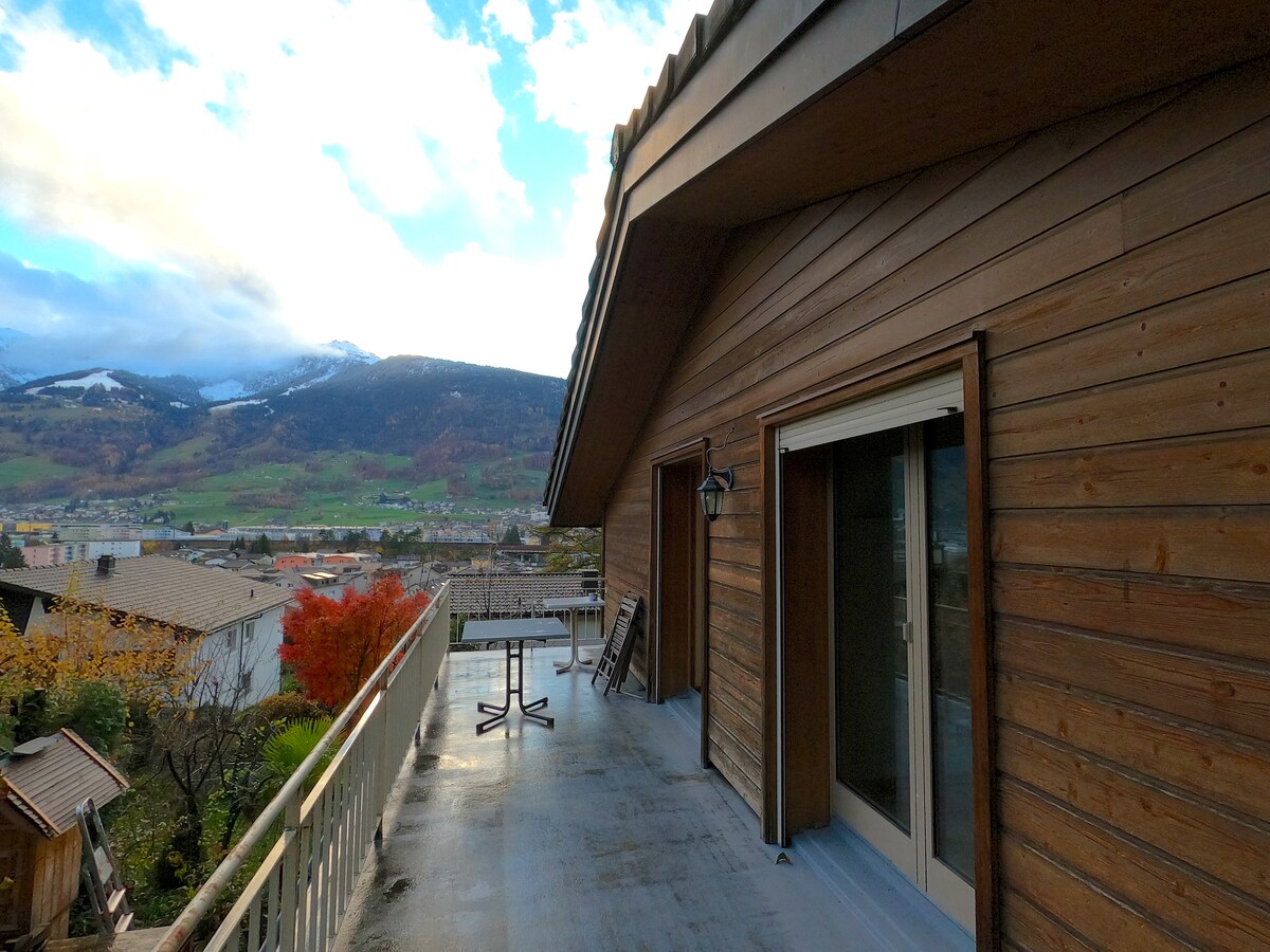 A spacious terrace is shown with wooden paneling and a railing overlooking a scenic valley and mountains. A small table and chairs are positioned against the wall, with lush greenery visible below and sky with clouds above.