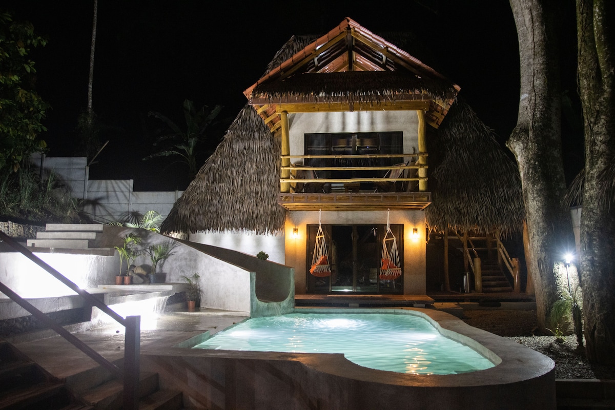 The villa’s exterior is illuminated at night, showcasing thatched roofs and balcony hammocks. A gently lit pool, framed by tropical landscaping, reflects a serene ambiance. Steps lead from the pool area to the entrance of the uniquely designed two-level structure.