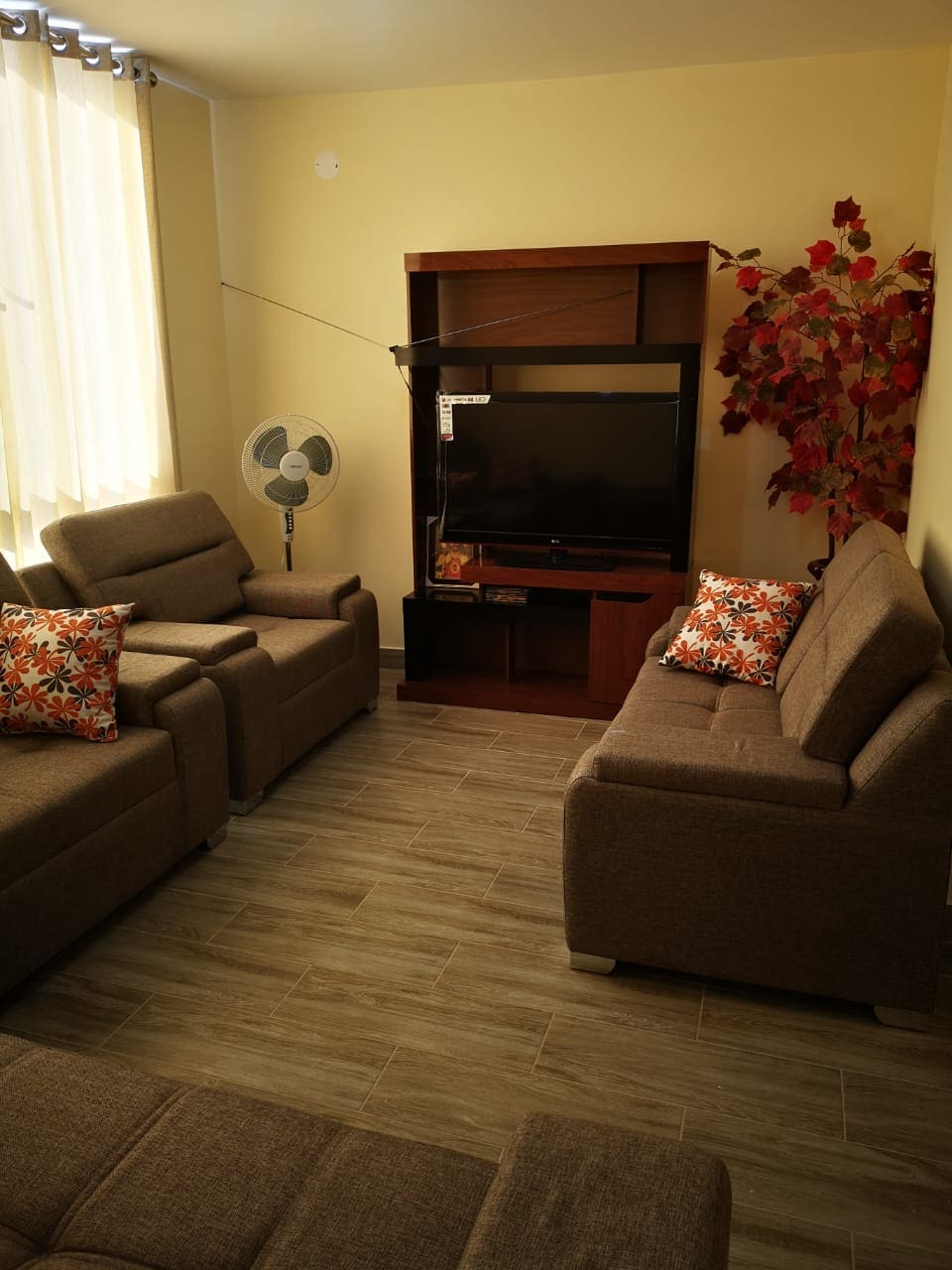 A seating area is presented with two comfortable armchairs and a spacious sofa, complemented by decorative throw pillows. A television is positioned on a wooden console, with a small fan nearby. Natural light illuminates the room through sheer window coverings.