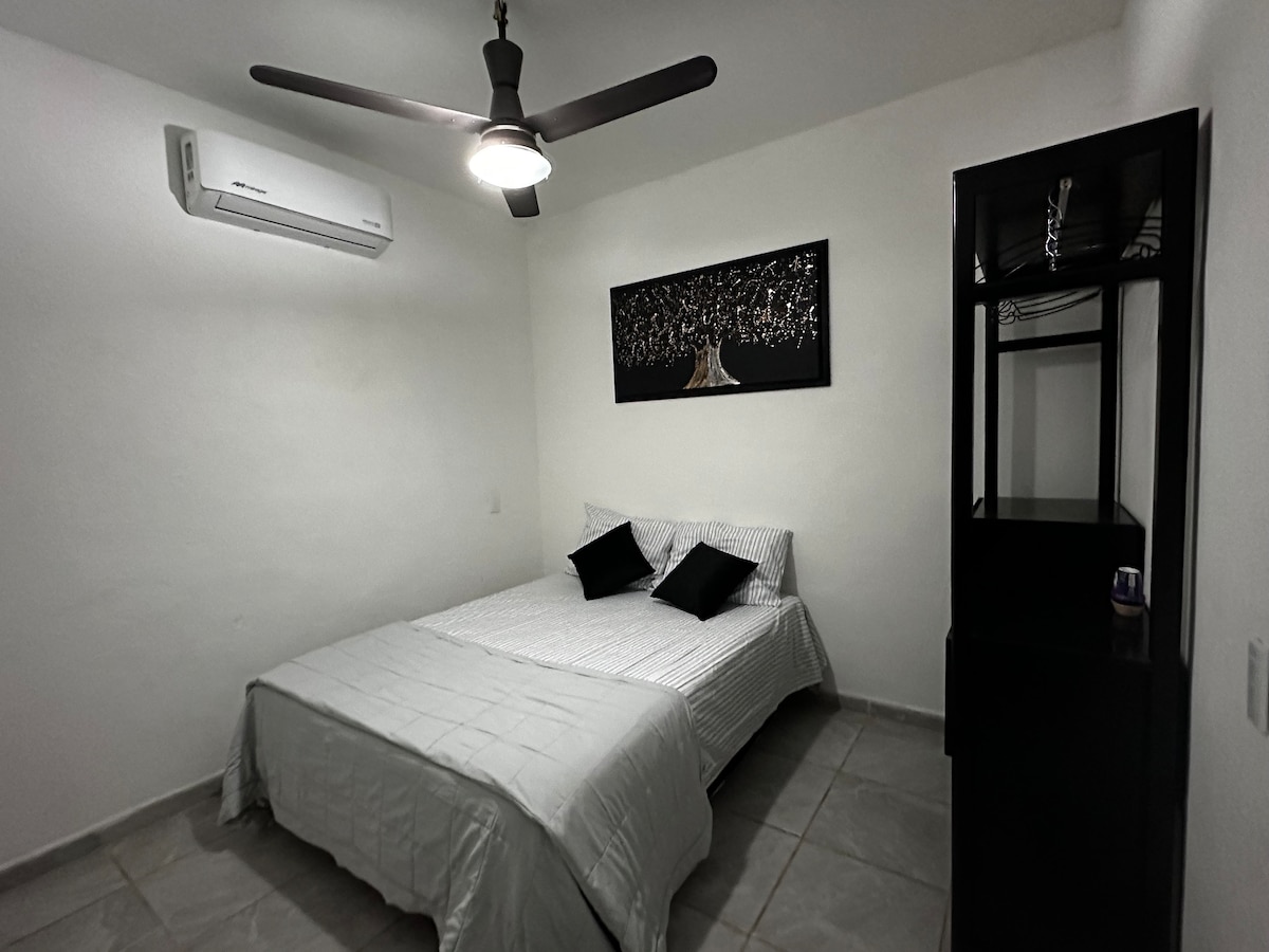 A spacious sleeping area is observed, featuring a comfortable bed with a light-colored coverlet and two black pillows. A black dresser and wall art complete the space, while a ceiling fan and air conditioning unit provide climate control.