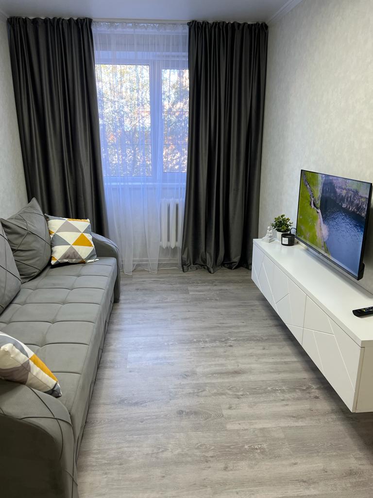 A cozy living area is presented, featuring a gray sofa adorned with a geometric cushion. A low white media console holds a television, while a large window draped with sheer curtains allows natural light to illuminate the space.