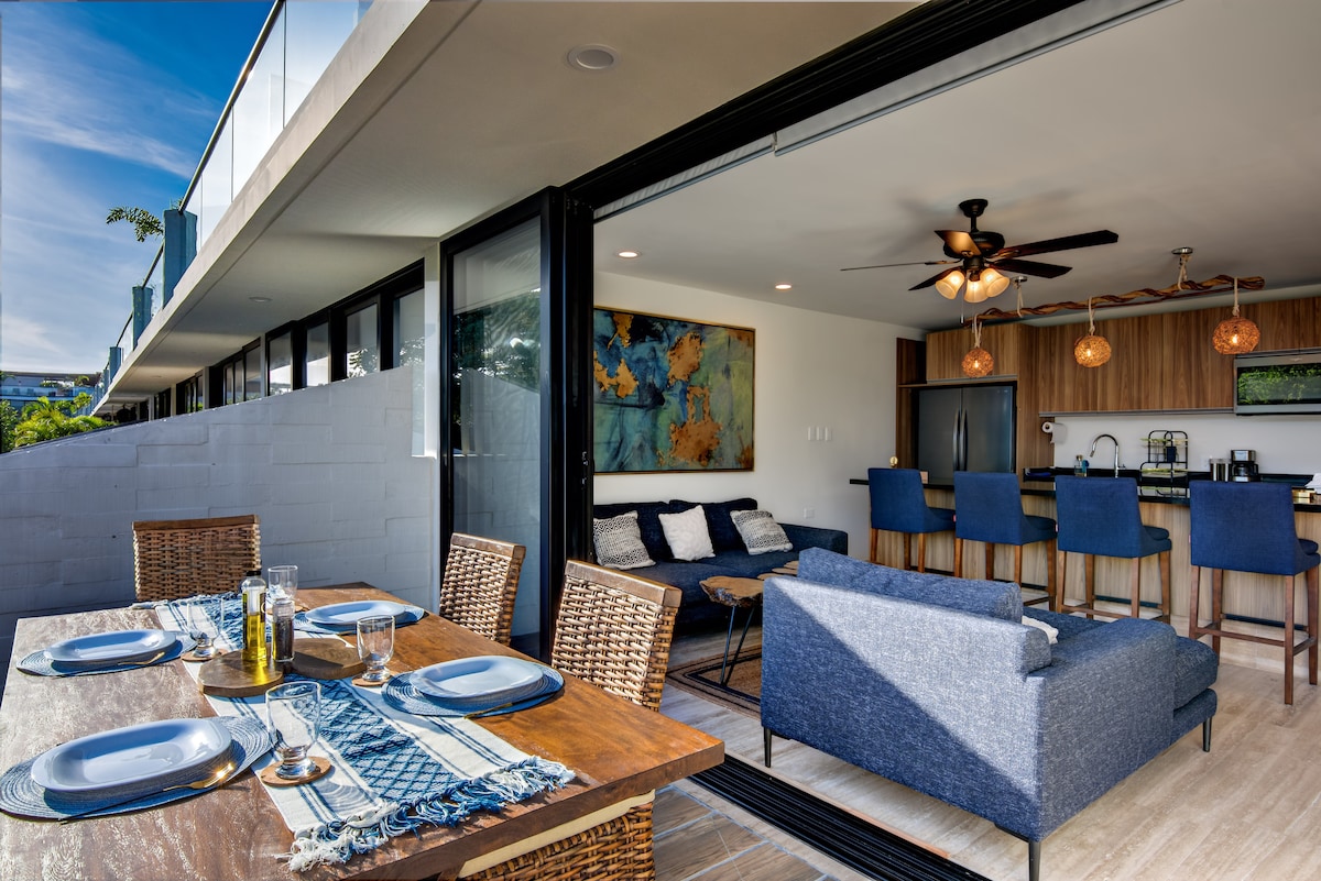 An open-concept living area is visible, featuring a modern kitchen with bar seating. A spacious dining table sets outside, complemented by woven chairs and table settings. Large glass doors connect the indoor space with the outdoors, allowing for natural light and a seamless transition.