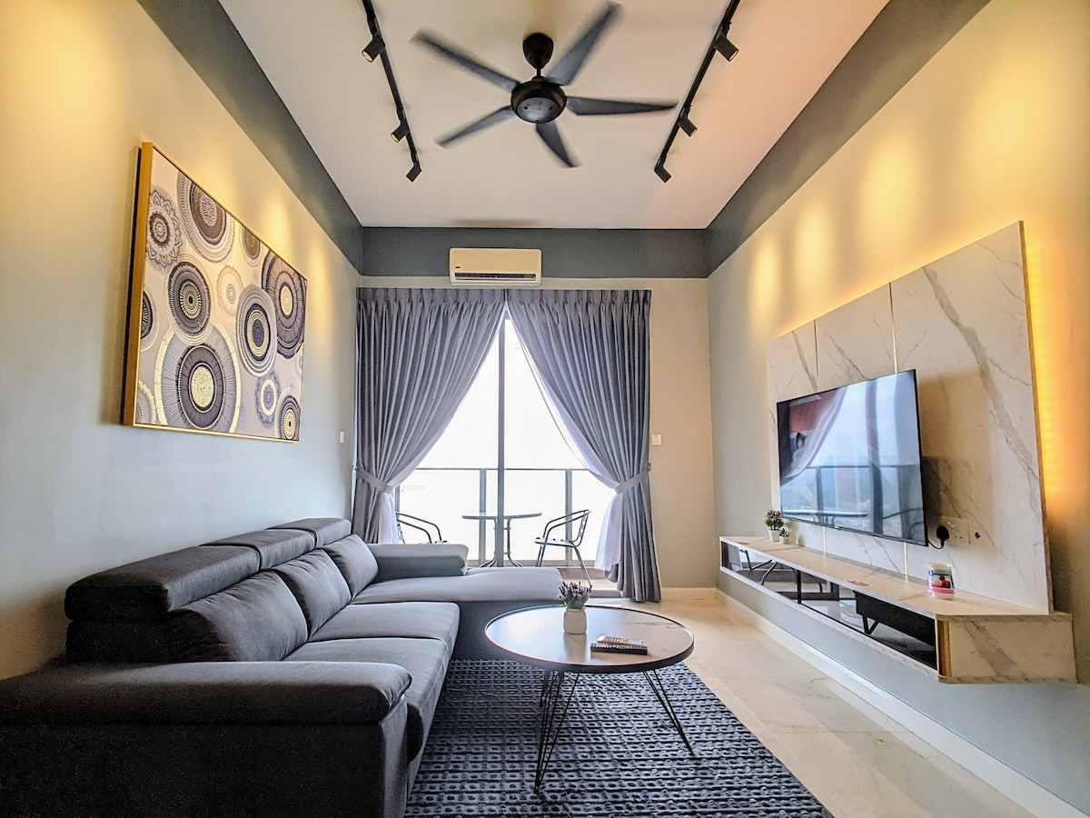 A modern living room features a dark grey sectional sofa arranged around a circular table. Floor-to-ceiling windows are dressed with elegant curtains, allowing natural light to enhance the space. A flat-screen television is mounted on a light marble panel, complemented by simple decorative elements.