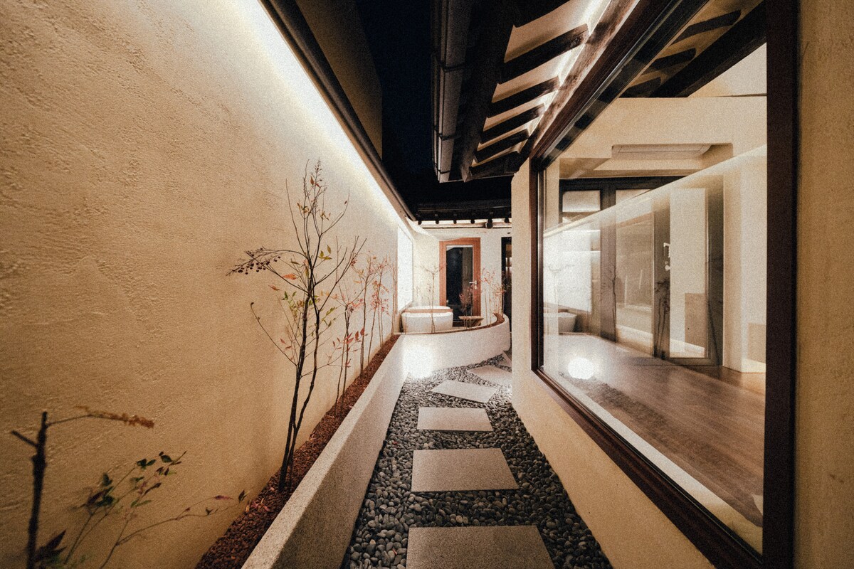 A serene pathway lined with smooth stepping stones leads through a well-lit outdoor space. Minimalist landscaping features decorative stones and subtle plant life, while a large window allows for views into the indoor area, enhancing the connection between nature and the interior.