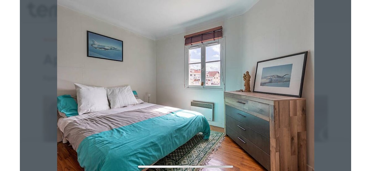 A bright bedroom features a spacious bed with a light blue duvet and white pillows. Natural light fills the room through a window adorned with a wooden blind, revealing a view of the surrounding area. A wooden dresser adds functionality and style against the light-toned walls.