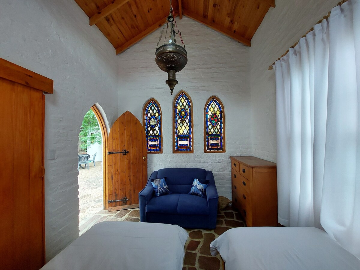 The room features high ceilings with exposed wooden beams, creating a spacious feel. Three stained glass windows provide vibrant color, illuminating the area. A blue couch is positioned between two twin beds, with natural light filtering through flowing white curtains.
