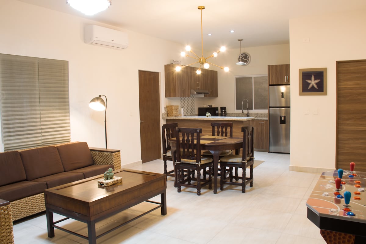A modern living area is presented with a comfortable brown sofa, a wooden coffee table, and a dining table for four. The well-equipped kitchen features modern appliances, and decorative accents like a starfish enhance the simple decor. Natural light enters through the windows with blinds.