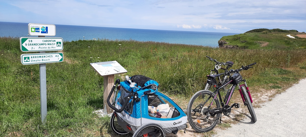 A scenic coastal path is visible with bicycles parked beside a child trailer. A signpost displays distances to various destinations, including Omaha Beach. The lush green landscape contrasts with the blue sea in the background, and a clear sky enhances the outdoor atmosphere.