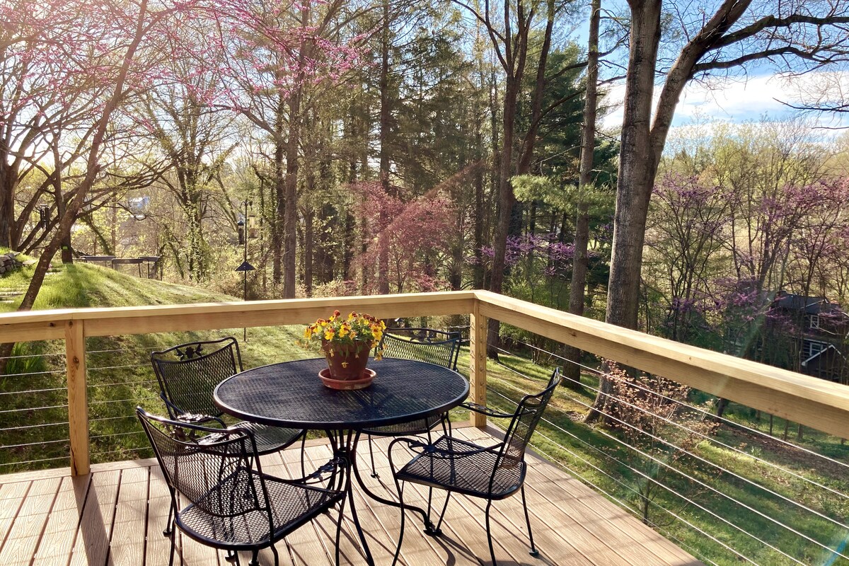 An outdoor deck features a wrought iron dining table surrounded by four matching chairs. A potted plant sits at the center of the table. Surrounding greenery and flowering trees create a serene backdrop, inviting relaxation in a peaceful setting.