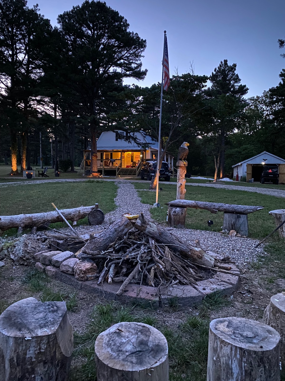 An outdoor gathering space features a fire pit surrounded by log stools. A stone pathway leads towards a house with illuminated porch lights, framed by tall trees. An American flag stands nearby, adding a touch of patriotism to the serene setting.