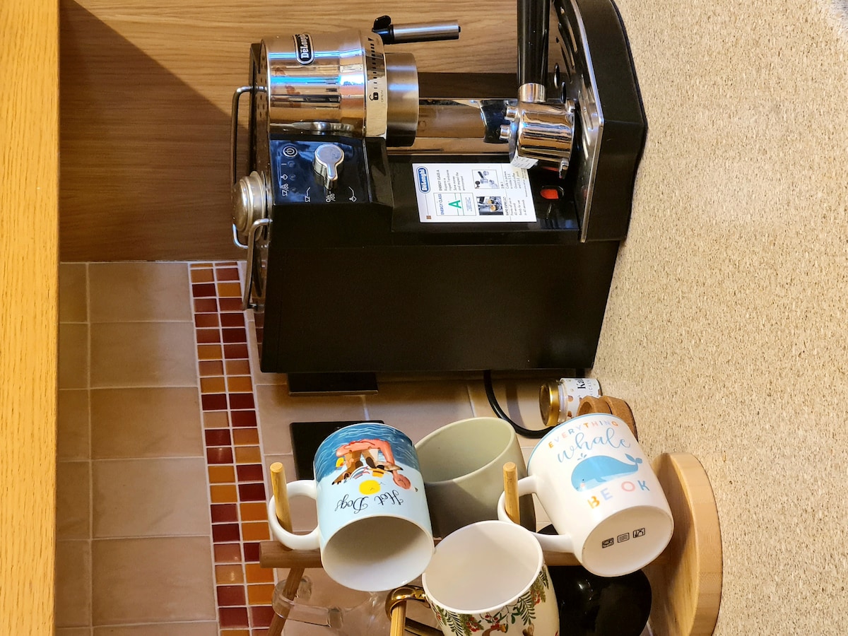 A coffee maker sits on a counter, accompanied by a selection of mugs arranged on a small wooden stand. The mugs display various designs and colors, creating a casual and functional coffee station.