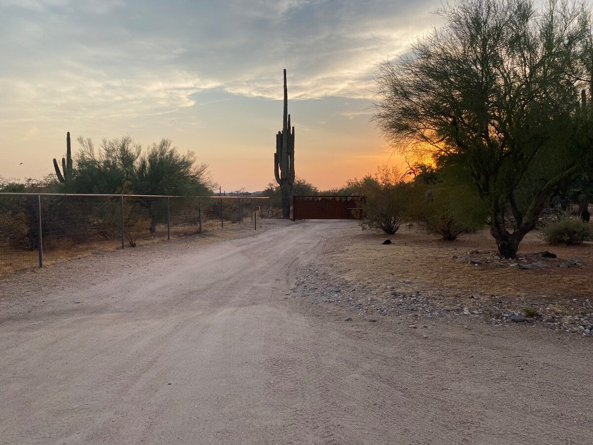 A gravel road leads to a fenced entrance, flanked by tall cacti and desert vegetation. The warm hues of a sunset illuminate the sky, creating a serene setting in the Arizona desert landscape.