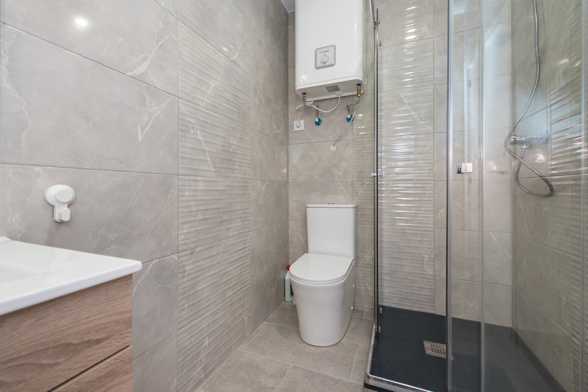 A modern bathroom features light gray tiled walls and flooring. A glass walk-in shower is present alongside a white toilet. A wooden vanity with a sink occupies one side, adding a touch of warmth to the sleek design.