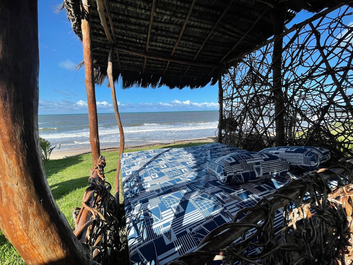 A shaded lounging area features a spacious bed adorned with patterned bedding, overlooking the beach. The natural structure, crafted from intertwined branches, provides a relaxing spot to enjoy coastal views and gentle sea breezes.