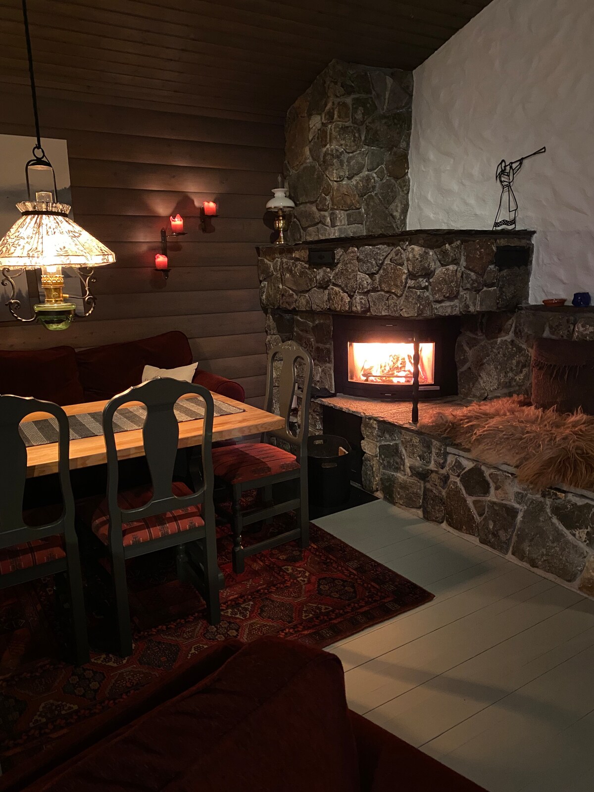 A cozy dining area is featured beside a stone fireplace, providing warmth and ambiance. The wooden table is flanked by dining chairs, and a plush red sofa is positioned nearby. Soft lighting from a decorative lamp enhances the inviting atmosphere.
