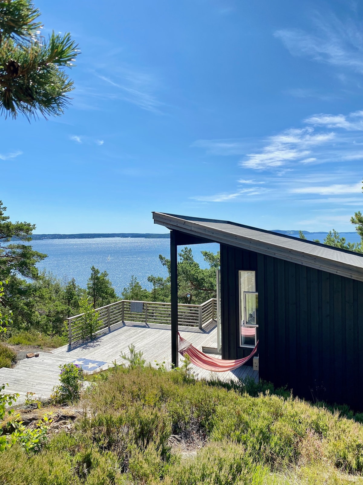 A sleek black cabin is shown alongside a spacious deck that overlooks the serene water. A hammock is suspended for relaxation under clear blue skies. Lush greenery frames the view, creating a peaceful outdoor environment.