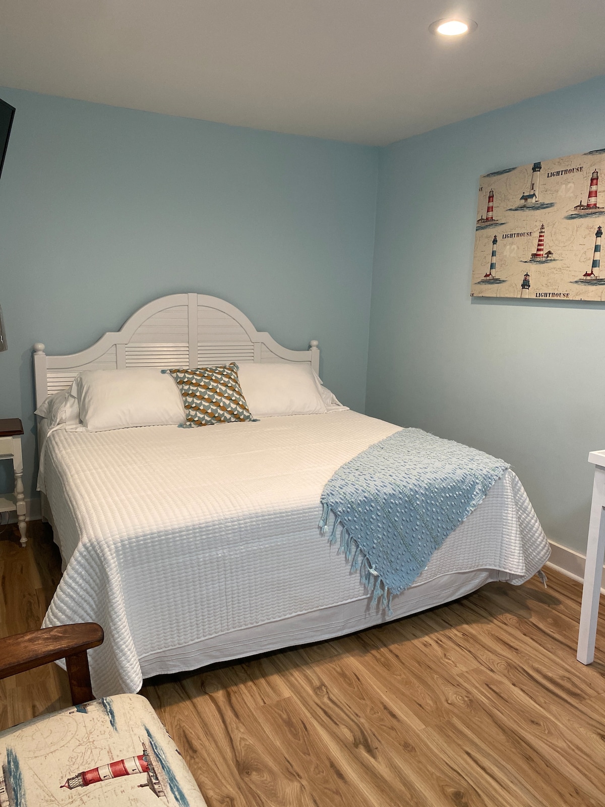 A well-appointed bedroom features a spacious bed with crisp white bedding and a decorative pillow. A light blue wall adds a calming backdrop, while a small accent quilt is draped over the bed. Natural wood flooring and a nautical-themed wall art piece enhance the cozy ambiance.