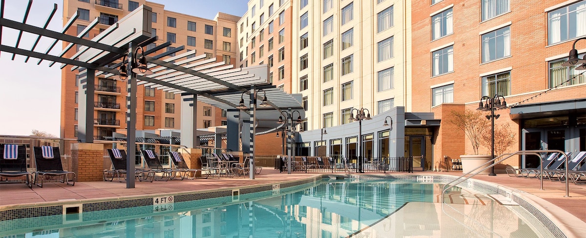 An outdoor pool area is accompanied by lounge chairs arranged under shaded pergolas. The surrounding brick buildings reflect a modern architectural style, with large windows framing the outdoor space. The pool features a clean, inviting design and a steps entryway.