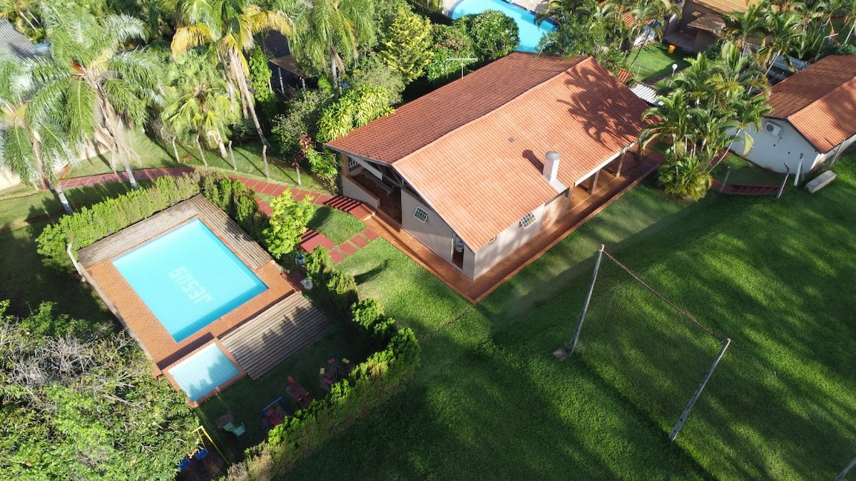 An aerial view of the property reveals a spacious outdoor area featuring a rectangular pool surrounded by wooden decking. Lush greenery and palm trees enhance the landscape, while a main house and additional structures are visible, emphasizing the inviting leisure spaces.