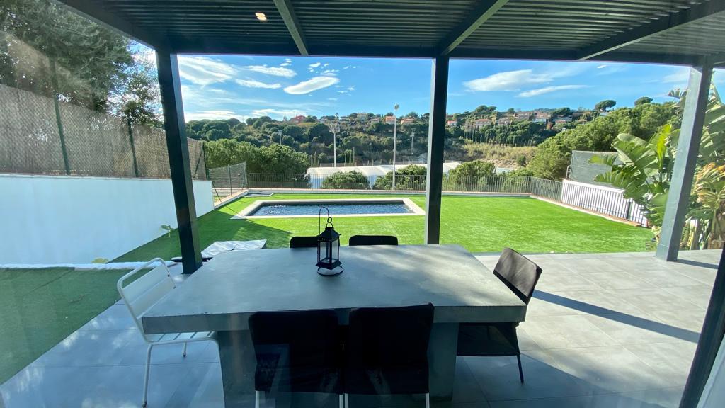 An outdoor dining area is visible, featuring a large table surrounded by black chairs. In the background, a swimming pool is situated amidst a serene landscape, with green grass and foliage enhancing the tranquil atmosphere. Clear skies complement the peaceful setting.