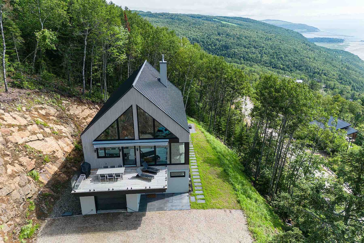 The chalet is set amidst a lush landscape, featuring a modern design with large glass windows that provide expansive views of the surroundings. A spacious outdoor patio is visible, equipped with seating, and the property is framed by trees that enhance its natural setting.