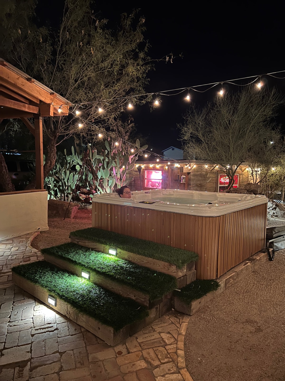 A hot tub is positioned at the base of illuminated steps, accented by soft green grass-like turf. String lights crisscross overhead, casting a warm glow over the inviting outdoor space, surrounded by desert vegetation and rustic stone pathways.