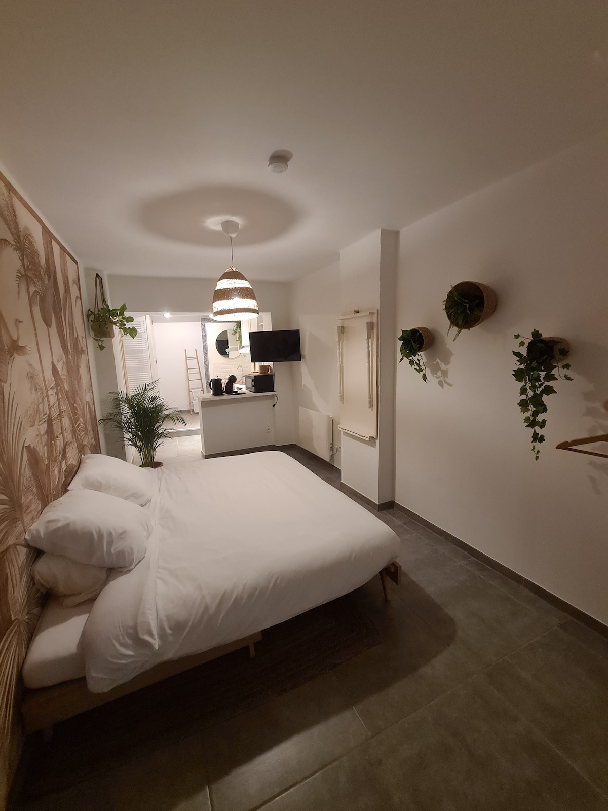 A spacious sleeping area features a comfortable bed with neatly arranged white bedding. The room is softly lit by a pendant light, and lush greenery is present near the walls. A kitchenette area is visible in the background, complemented by a small seating arrangement.