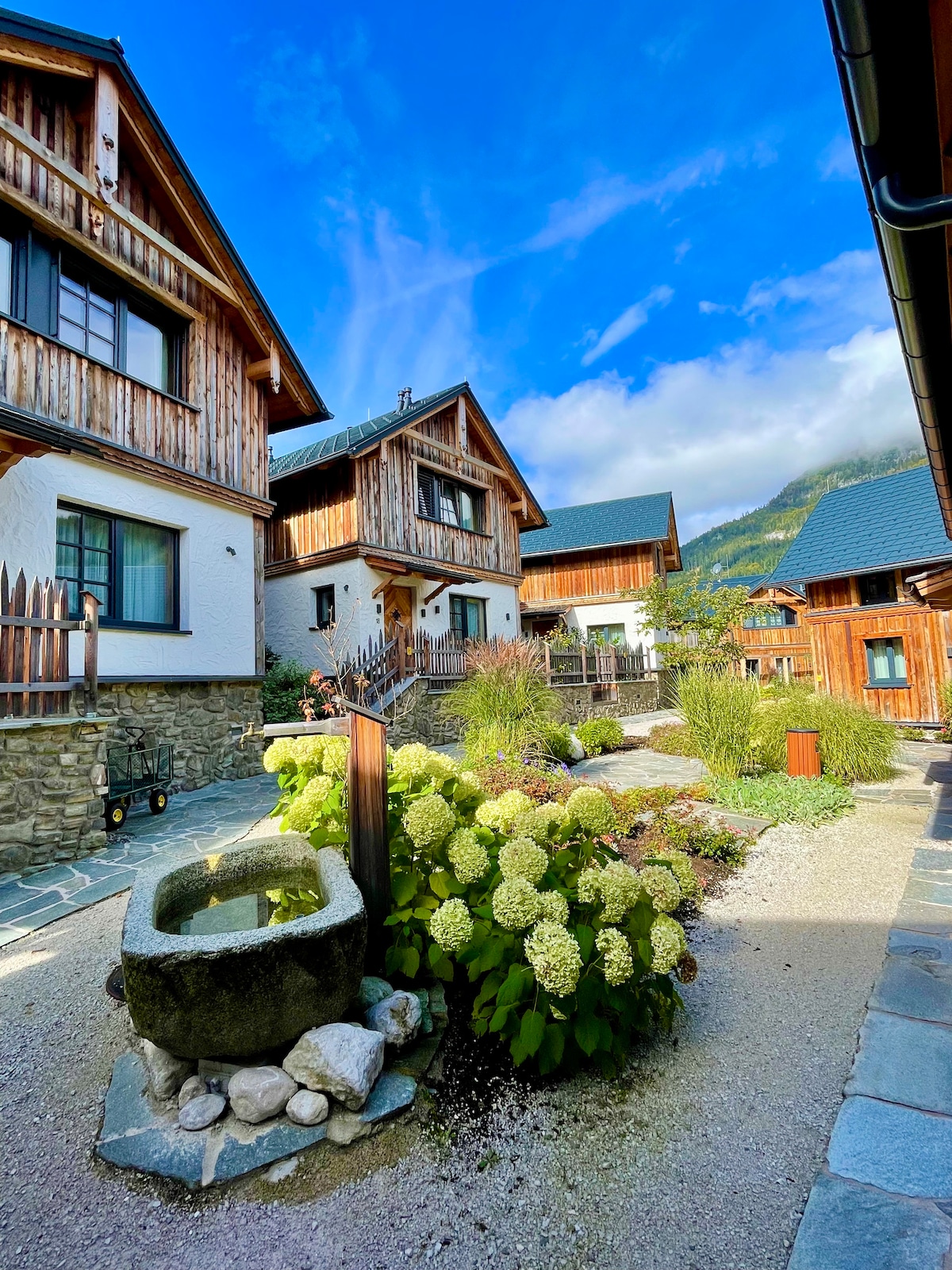 A charming courtyard surrounds the property, featuring a decorative well and lush hydrangea bushes. The rustic wooden buildings offer a blend of traditional and modern architecture under a clear blue sky, with greenery enhancing the inviting atmosphere.
