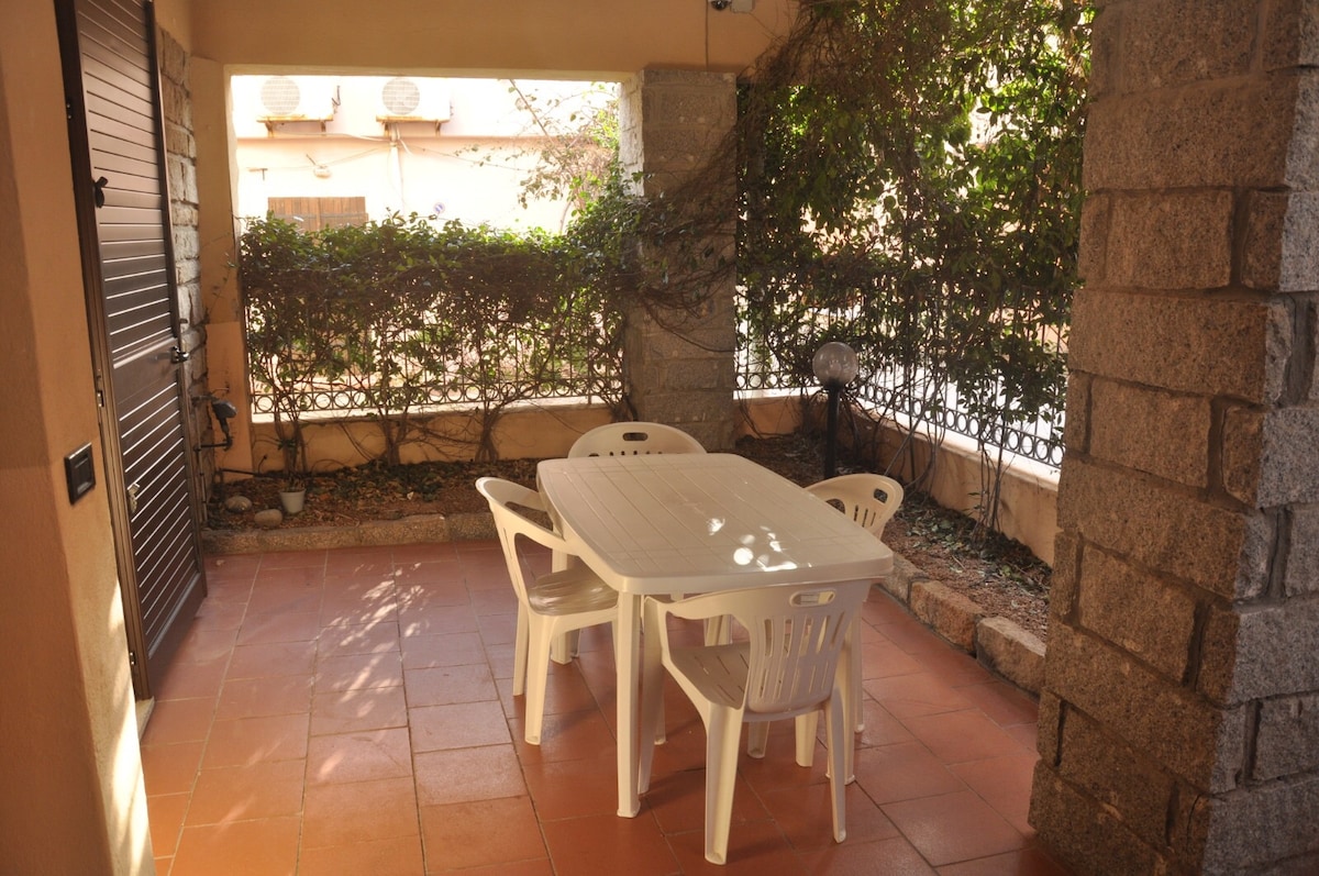 A covered outdoor area features a rectangular table surrounded by four white chairs. Lush greenery is visible in the background, providing a natural setting. The earth-toned tiled flooring complements the warm ambiance of the space.