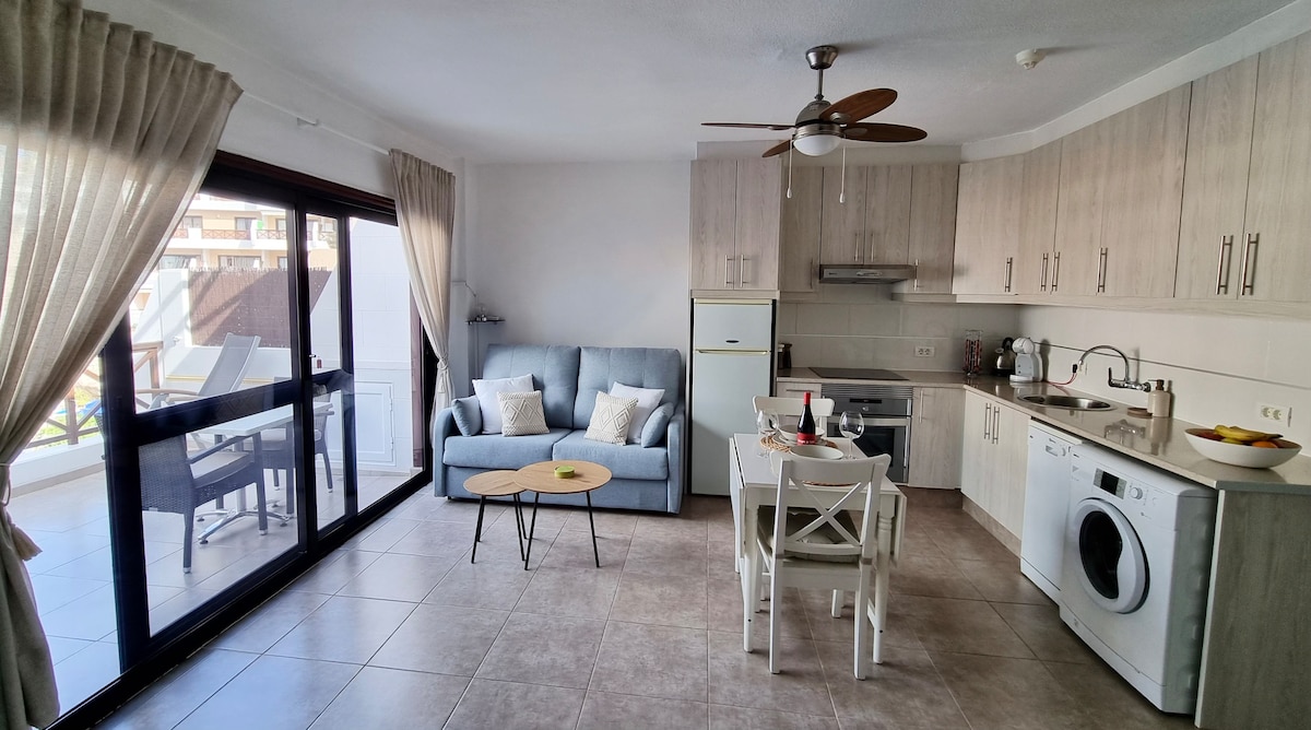 A modern open-plan living area features a light blue sofa, dining table, and a well-equipped kitchen. Large glass doors provide access to an outdoor space. Soft natural light enters through the curtains, and a washing machine is positioned near the kitchen for convenience.