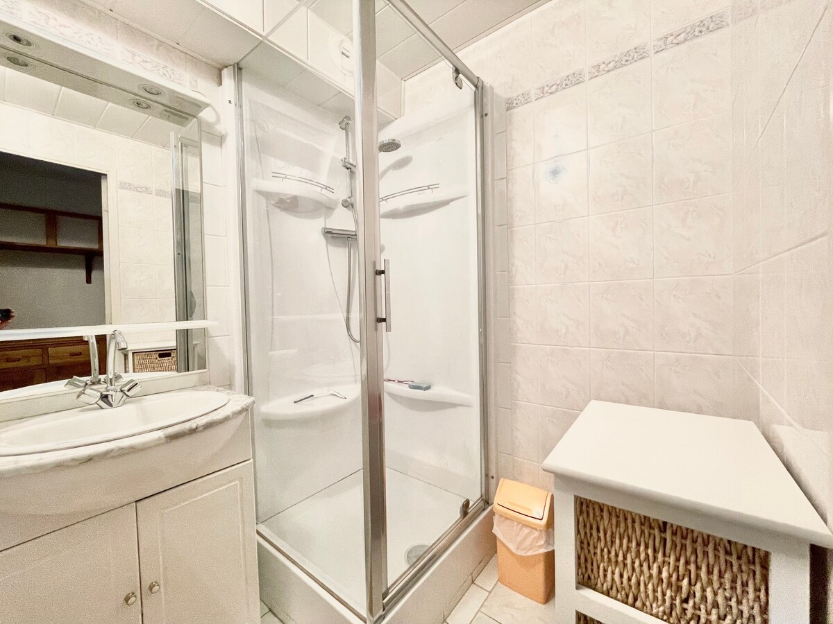 A bathroom features a shower enclosure with clear glass doors, allowing visibility of dual shower heads. A light-colored vanity with a marble countertop and a round sink is positioned beside the shower. A wicker basket and a trash can are included nearby.