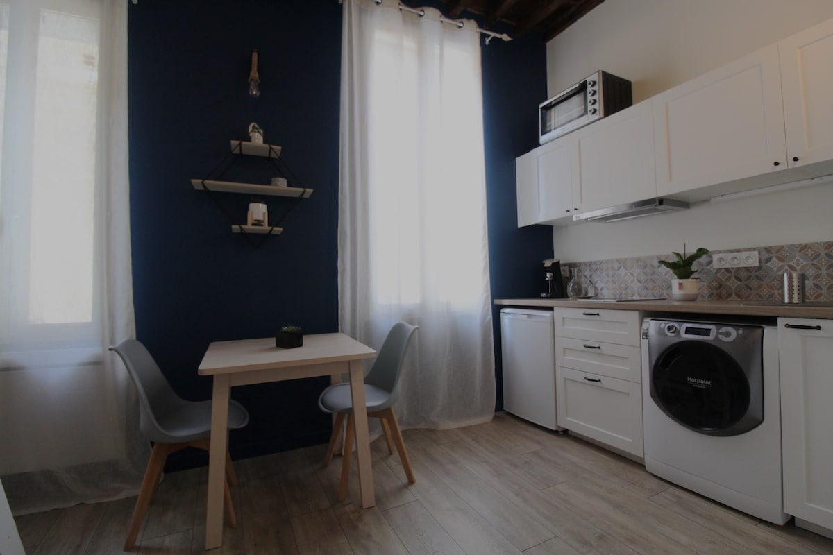 The kitchen features a compact dining area with a small table and two chairs. A white cabinetry and appliances are complemented by a dark blue accent wall. Light enters through two windows, while a shelf displays decorative items and a plant.
