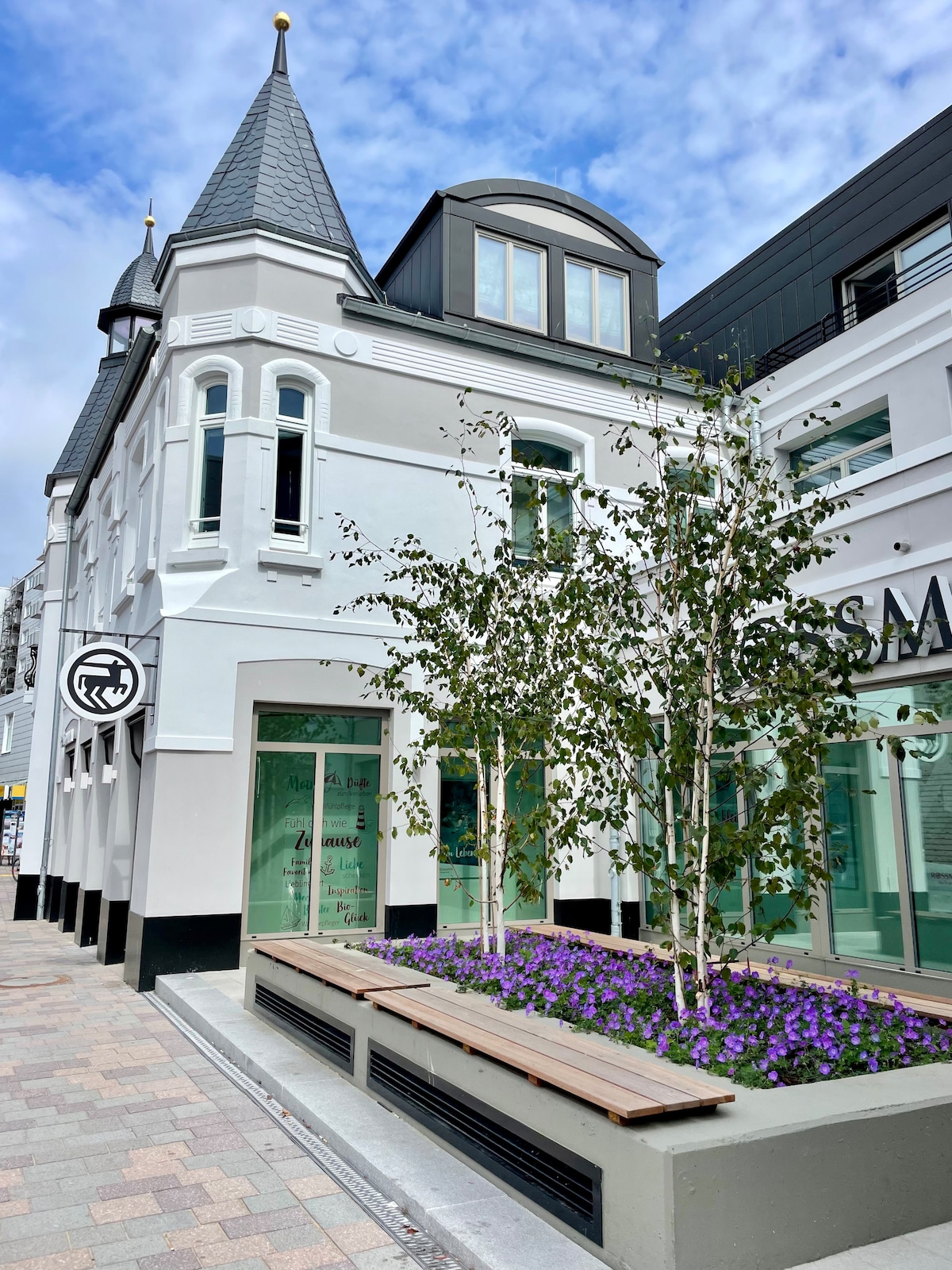 The exterior of a beautifully renovated historic building features a unique architectural design with pointed roofs. Large windows allow for ample natural light. Flower beds with purple blooms and slender trees enhance the entrance, providing a pleasant aesthetic contrast to the stone façade.