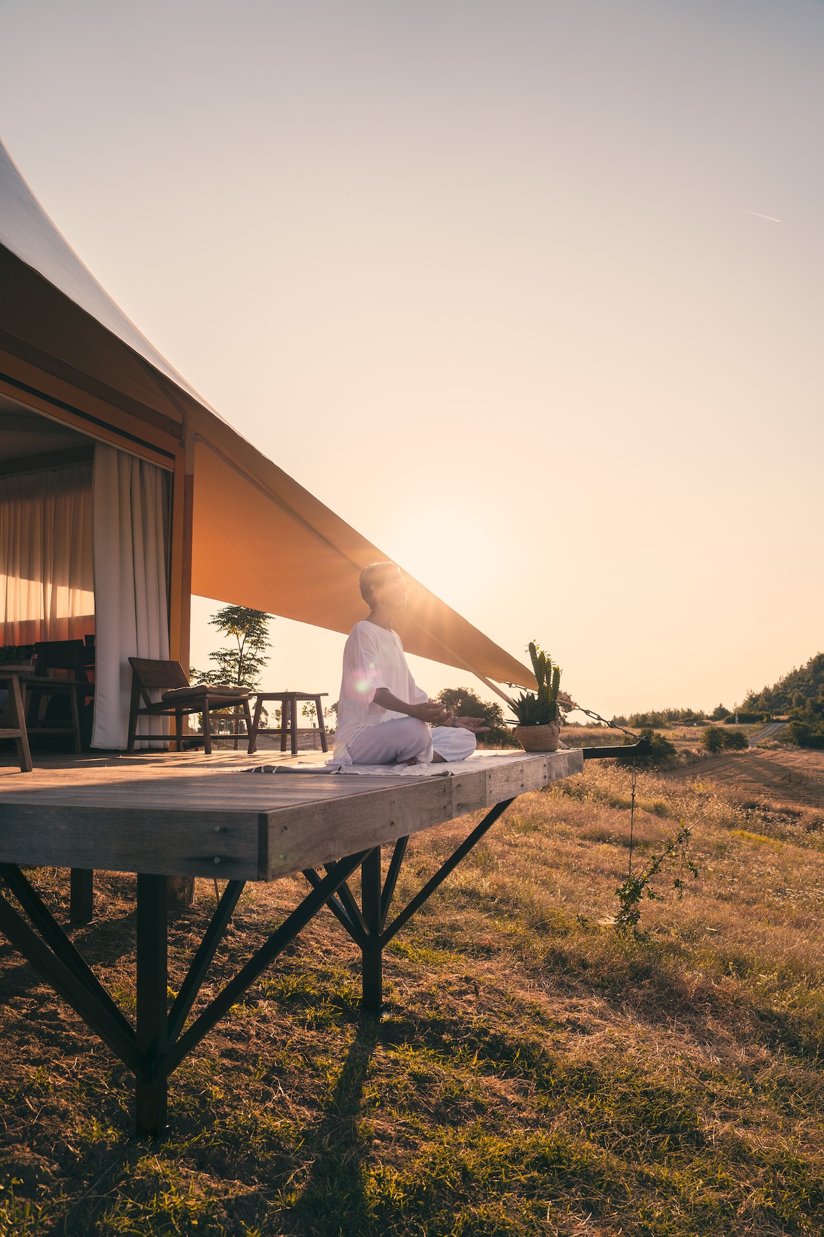 A luxurious tent is shown on a wooden deck, with a guest seated in meditation facing the sunrise. Natural light filters through the tent's soft fabric, highlighting the serene environment. Surrounding greenery and a hint of distant hills complete the peaceful setting.