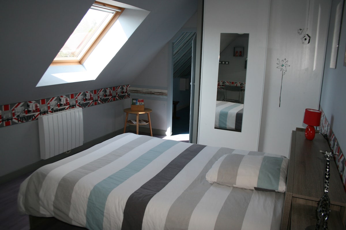 A well-lit bedroom features a spacious bed dressed in striped bedding. A narrow mirror reflects the room, while a small chair sits beside the bed. Natural light enters through a skylight, highlighting the walls decorated with a stylized border.