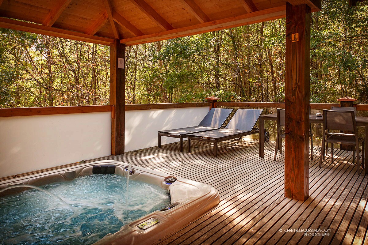 A wooden deck features a hot tub with water gently swirling. Two sun loungers are positioned nearby, and a small table accompanies a chair for relaxation. Lush greenery surrounds the area, providing a serene and private atmosphere.